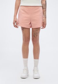 Shorts rose clair en tissu doux, dotés d'une ceinture élastique et d'un ourlet roulé. Portés avec des chaussettes blanches et des baskets.
