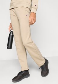 Μπεζ παντελόνια φόρμας με ελαστική μέση και πλαϊνές τσέπες. Μαύρα αθλητικά παπούτσια με υφή από δίχτυ. Μαύρο μπουκάλι νερού που κρατείται στο χέρι.