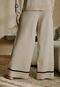 Beežid laia säärega püksid, mis on tehtud pehmest kangast, millel on must ääris ääres ja lame vöökoht, nähtud tagantpoolt.