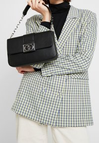 Woman in checkered blazer and turtleneck holding a black leather handbag with silver chain strap and decorative clasp.