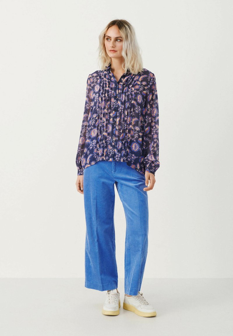 Luźna bluzka w granatowym kolorze z wzorem w fioletowe, pomarańczowe i białe motywy kwiatowe. Połączona z jasnoniebieskimi spodniami z korduroy i białymi trampkami.