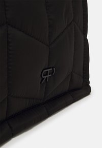 Section de tissu matelassé noir avec une couture géométrique subtile et un petit logo noir en relief près du coin inférieur sur un fond clair.