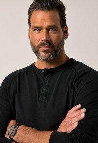 Hombre con cabello oscuro y barba, vistiendo una camiseta negra de tipo henley y un reloj de muñeca, de pie con los brazos cruzados, mirando con confianza hacia adelante.