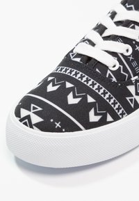 Baskets en toile noires avec un motif géométrique blanc, lacets plats blancs et une semelle extérieure en caoutchouc texturé blanche.