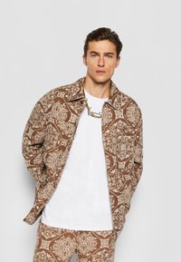 Brun jakke med mønster og intrikate cremede designs, hvid t-shirt under, og en guld kædede halskæde. Casual pasform med krave.