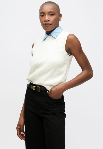 Mujer con la cabeza rapada que lleva un chaleco de punto blanco sin mangas sobre una camisa de color azul, pantalones negros y un cinturón negro, de pie con una mano en el bolsillo.