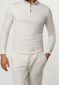 Vaaleabeige pitkähiha-pikeepaita, jossa on kaulus ja kolme nappia. Pehmeästi teksturoitu kangas ja istuva malli. Yhdistetty vastaaviin verkkareihin.