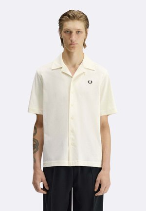 Giovane uomo con capelli effetto bagnato che indossa una camicia crema a maniche corte con bottoni e un logo ricamato nero sul petto, accompagnata da pantaloni neri.