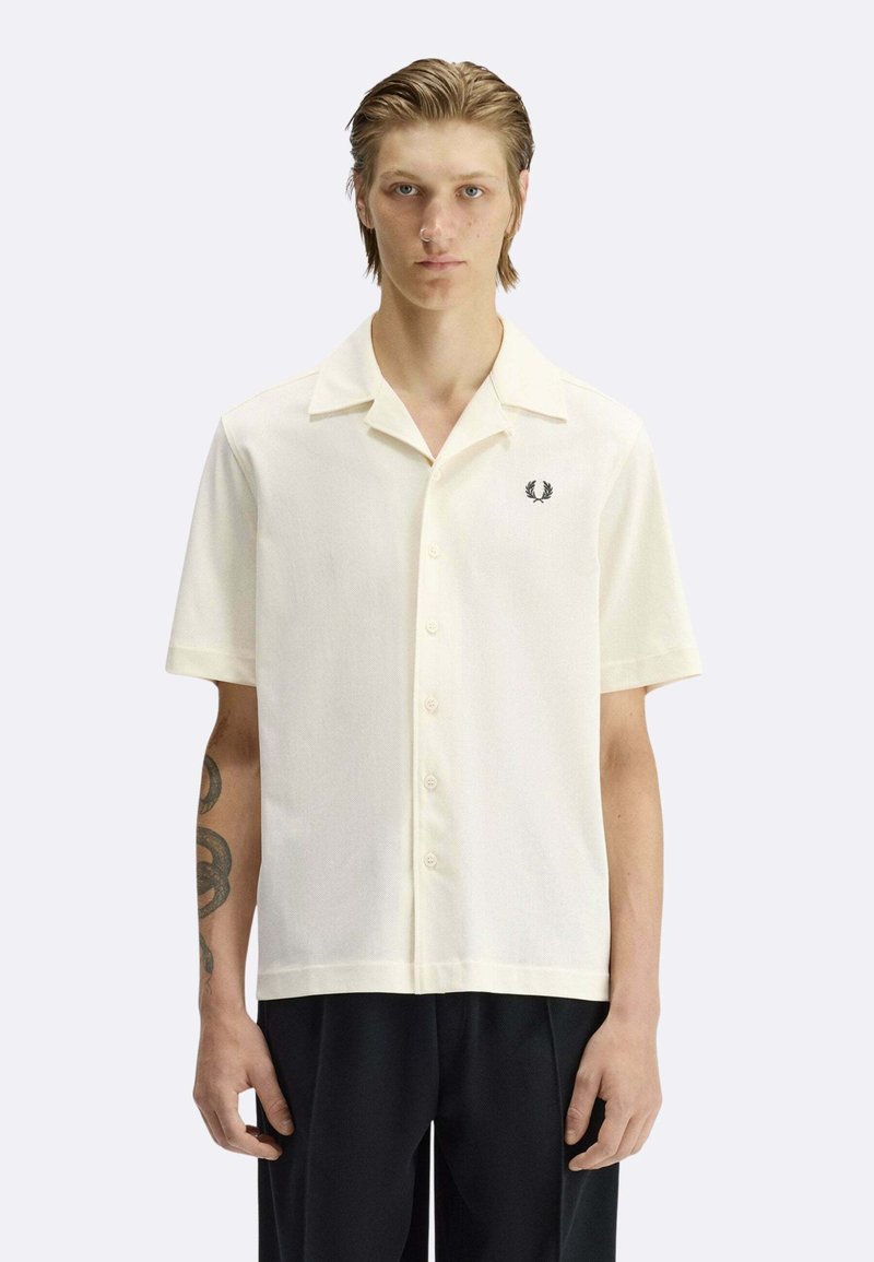 Jeune homme aux cheveux mouillés portant une chemise crème à manches courtes avec un logo brodé noir sur la poitrine et un pantalon noir.
