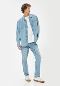 Camicia di denim azzurro chiaro con bottoni frontali, tasca applicata e jeans azzurri chiari abbinati. T-shirt bianca sotto e sneakers beige con suole marroni.