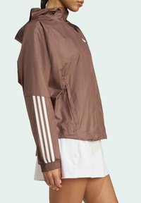 Braune leichte Jacke mit hohem Kragen, mit weißen Streifen an den Ärmeln und elastischen Bündchen. Inklusive seitlicher Taschen.