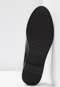 Semelle noire d'une chaussure habillée avec talon à la cheville, motif texturé, et nom de la marque "ANNA FIELD" visible sur la semelle.