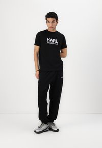 Μαύρη μπλούζα με άσπρο λογότυπο "KARL LAGERFELD", σε συνδυασμό με μαύρες φόρμες και ασημένια αθλητικά παπούτσια, όλα σε απλό λευκό φόντο.