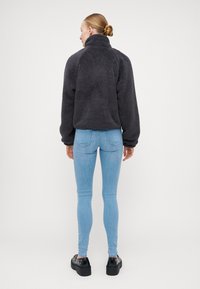 Fleece jakna v temno sivi barvi z visokim ovratnikom in oversized rokavi, v kombinaciji s svetlo modrimi ozkimi džinsom in črnimi debelimi platform čevlji.