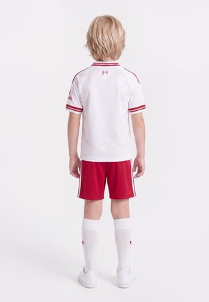 Małe dziecko stojące tyłem, ubrane w biały sportowy jersey z czerwonymi paskami, czerwone szorty, białe skarpetki do kolan i białe buty.