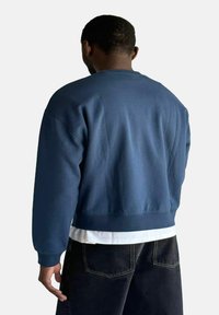 Felpa blu navy con collo a giro e orlo a coste. Il tessuto appare liscio con una vestibilità comoda. Indossata sopra un top bianco a strati e pantaloni scuri.