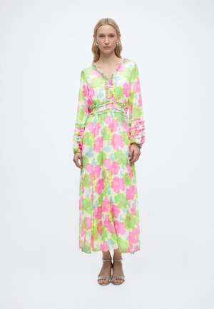 Kvinne iført en lang blomstrete kjole med rosa, grønne og blå mønstre, står rett opp med hendene langs sidene på hvit bakgrunn.