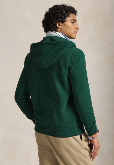 Camisola de capuz verde com textura suave, com capuz forrado e punhos canelados, combinada com calças bege. Design simples sem padrões visíveis.