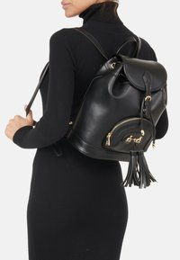 Musta nahkareppu pyöreällä muodolla, kultaisilla detaljeilla, vetoketjullisella taskulla ja tupsuaksentilla. Suora pinta, nauhakiinnitys.