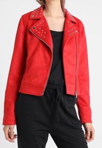 Veste de motard en suède synthétique rouge avec des clous en argent sur le col. Dispose d'une fermeture éclair et d'une silhouette ajustée avec une texture lisse.