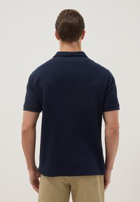 Hombre vistiendo una camisa polo de manga corta azul marino y pantalones beige, visto desde atrás contra un fondo claro y liso.