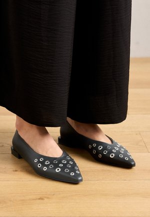 Black leather flats with a pointed toe, featuring metal eyelet accents in varying sizes, and a low square heel. Smooth texture on wooden floor.