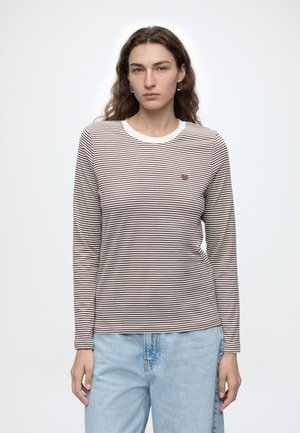 Jeune femme aux cheveux bruns et raides portant un t-shirt à manches longues rayé blanc et marron et un jean bleu clair sur un fond blanc uni.