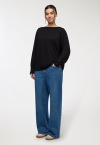 Czarny sweter z długim rękawem i okrągłym dekoltem, z guzikami na rękawach. Połączony z niebieskimi dżinsami w szerokim kroju i beżowymi butami.