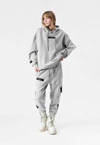 Sudadera gris con corte oversize, cordones y detalles en etiqueta negra, combinada con pantalones jogger a juego con parches desgastados y cremalleras laterales.