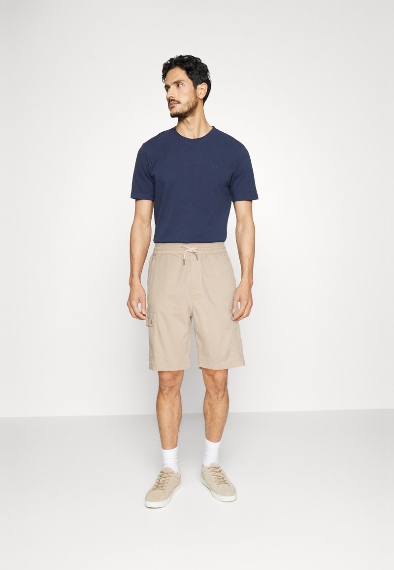 Homme portant un t-shirt bleu marine, un short cargo beige avec cordon de serrage, des chaussettes blanches et des baskets beiges, se tenant contre un fond blanc.