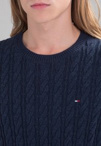 Maglione blu navy con lavorazione a treccia, realizzato in filato morbido. Presenta un collo rotondo e un piccolo logo rosso, bianco e blu sul petto.