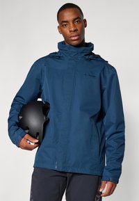 Jūras flotes ūdensizturīgā jaka ar priekšējo rāvējslēdzēju, regulējamu kapuci un elastīgiem piedurkņu galiem; modele tur melnu ķiveri pret gaišu fonu.