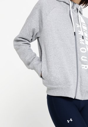 Femme portant un sweat à capuche zippé gris clair avec des cordons blancs et un legging bleu marine avec un petit logo Under Armour.
