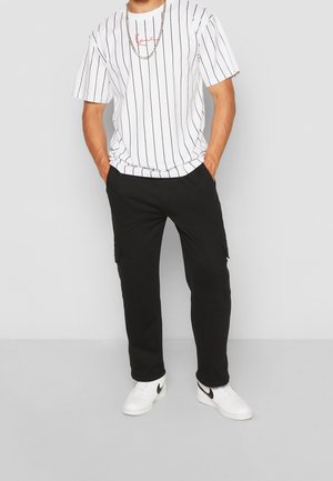 Weißes gestreiftes T-Shirt mit rotem Logo, kombiniert mit schwarzen Cargo-Hosen. Das Modell trägt weiße Sneaker mit schwarzen Akzenten. Lässiges Outfit-Design.