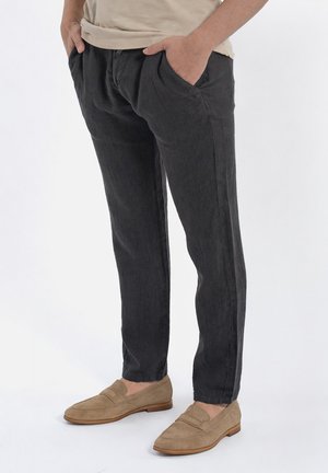 Persona che indossa pantaloni casual neri e mocassini beige in pelle scamosciata, in piedi con le mani in tasca su sfondo bianco.