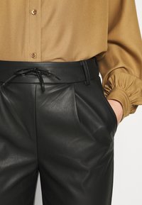 Svarta läderbyxor med veck och dragsko i midjan. Kombinerade med en ljusbrun blus med knappryck och samlade ärmar.