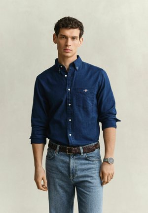 Jeune homme portant une chemise bleu foncé boutonnée aux manches retroussées, un jean bleu clair, une ceinture marron et une montre-bracelet argentée, debout devant un fond uni.