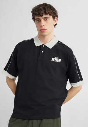 Giovane uomo con capelli ricci che indossa una polo nera Nike con colletto e polsini bianchi, in piedi davanti a uno sfondo chiaro e uniforme.