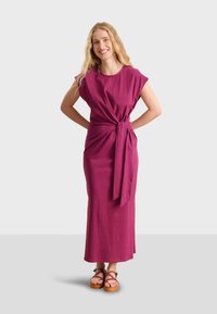 Fuchsia long dress with short sleeves, wrap style and knot detail at waist. Smooth fabric and ankle-length, paired with brown platform sandals.