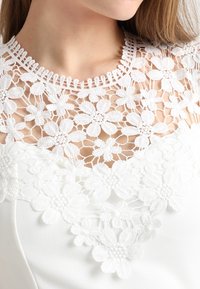Gros plan sur un décolleté en dentelle florale blanche porté par une personne à la peau claire et aux longs cheveux châtain clair.
