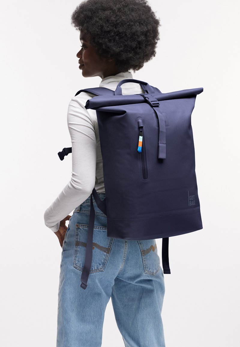 Persona con capelli corti e ricci che indossa una camicia bianca a maniche lunghe e jeans blu, che porta uno zaino grande blu navy con chiusura a rotolo.