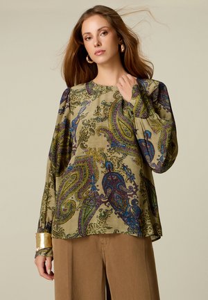 Mujer con cabello largo y castaño que lleva pendientes de aro dorados, una blusa de manga larga con diseño de cachemira y pantalones marrón claro sobre un fondo neutro.
