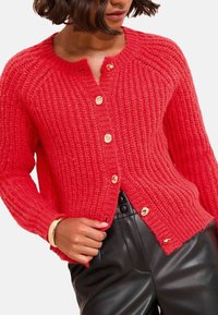 Cardigan a coste rossa con collo rotondo e bottoni in tono oro. Indossato sopra pantaloni in pelle nera, con una vestibilità comoda e superficie texture.