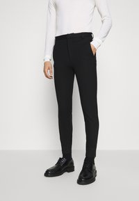 Pantalones negros ajustados con pierna cónica, con cinturón, bolsillos laterales y cremalleras en los tobillos. Combinados con una camisa blanca de manga larga y zapatos negros.
