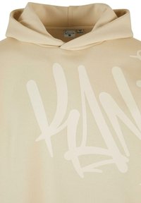 Sudadera beige con una textura suave y un gran diseño impreso en blanco en el frente. Cuenta con capucha con cordón y un corte relajado.