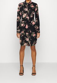 Abito chemisier nero a fiori con maniche lunghe, motivo di rose rosa e foglie verdi, vita annodata, chiusura con bottoni e colletto. Tacchi neri con cinturini.