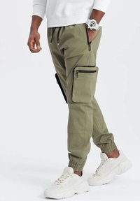Pantalons cargo vert olive avec poignets élastiques, poches zippées et poche cargo latérale avec bord noir, associés à des baskets blanches.