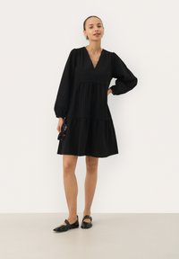 Robe noire longueur genoux avec de longues manches bouffantes, tissu texturé, col en V et jupe à volants. Le modèle tient des lunettes de soleil noires, se tenant sur un fond uni.