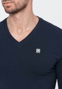 Laivan sininen V-neule t-paita, joka on valmistettu puuvillasta ja siinä on muotoiltu malli. Vasemmassa rintataskussa on valkoinen logo. Sileä pinta.