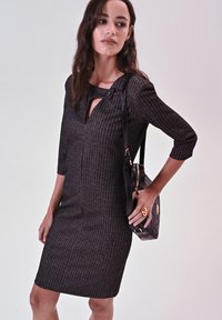 Graues, strukturiertes Kleid mit dreiviertellangen Ärmeln und einem Rundhalsausschnitt mit einem Schlitz. Das Modell trägt eine schwarze Handtasche mit goldenem Akzent.